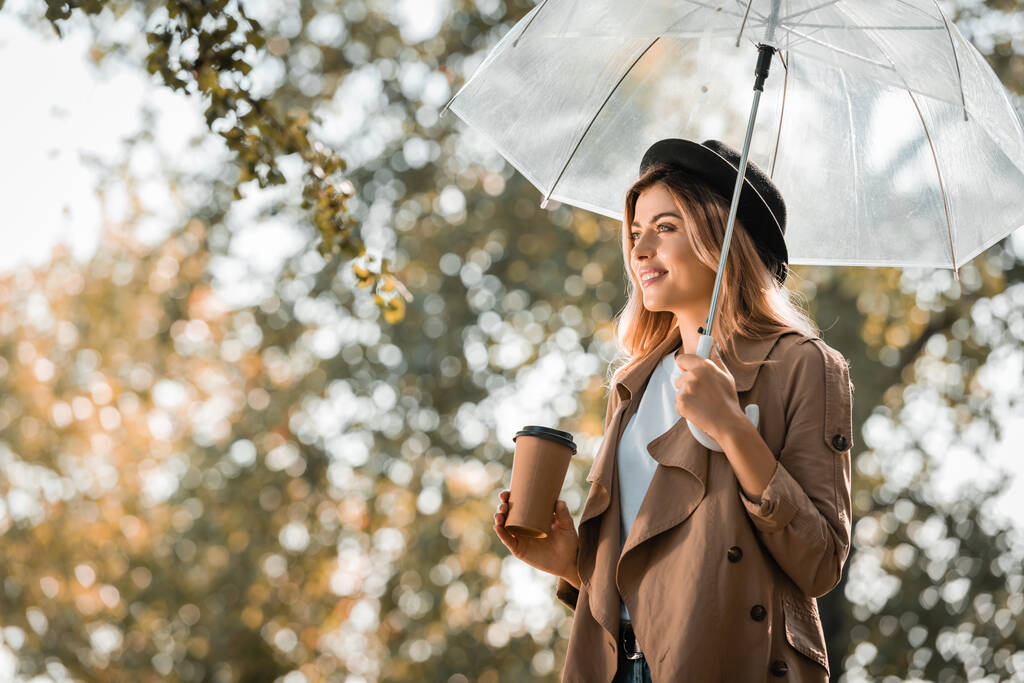 žena v trenčové bundě a klobouku stojící pod deštníkem a držící papírový kelímek v podzimním parku  - Fotografie, Obrázek