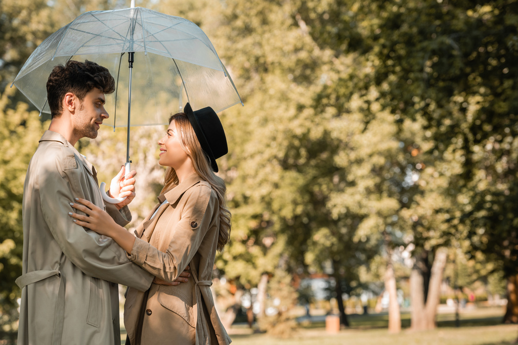 Seitenansicht einer Frau mit Hut und eines Mannes im Trenchcoat, die unter einem Regenschirm im herbstlichen Park stehen  - Foto, Bild