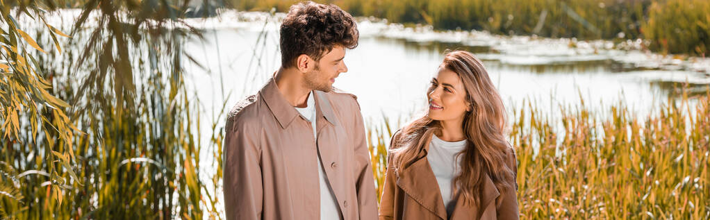 panoramiczne ujęcie zadowolonej kobiety i mężczyzny w płaszczach okopów patrzących na siebie nad jeziorem w jesiennym parku - Zdjęcie, obraz