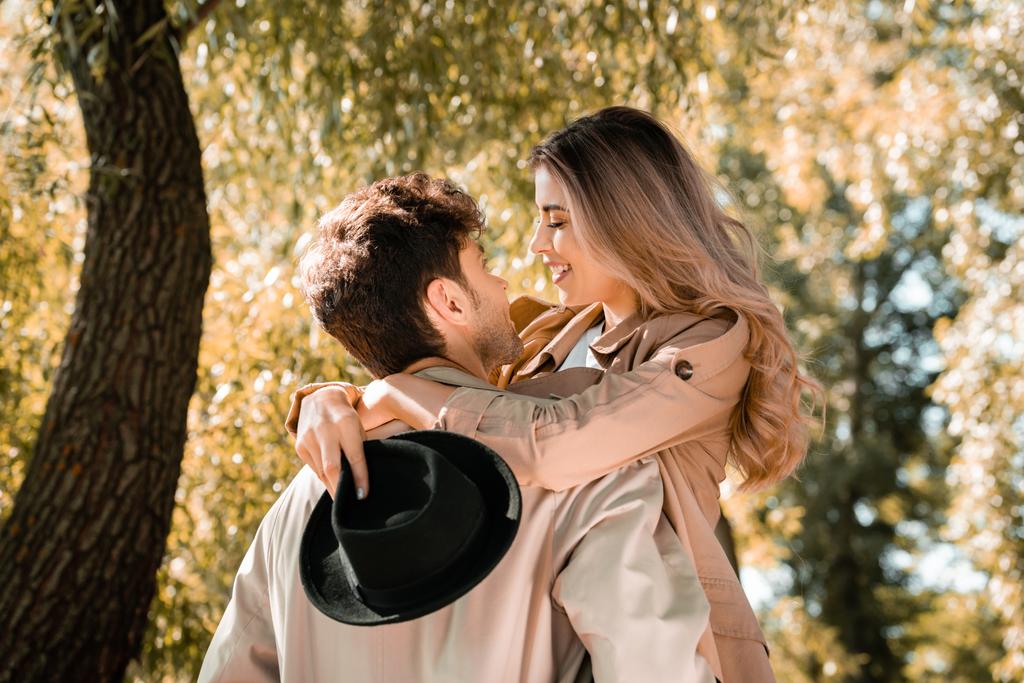 excited woman with hat hugging and looking at boyfriend in autumnal park - Photo, Image