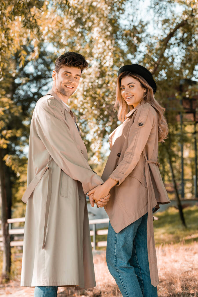 muž a žena v trenč kabáty při pohledu na kameru a držení se za ruce v podzimním parku  - Fotografie, Obrázek