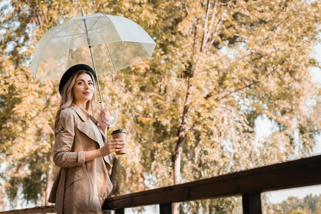 秋の公園に行くために傘とコーヒーと使い捨てカップを持って帽子の女性  - 写真・画像