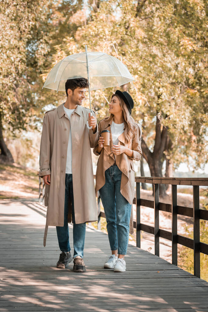 casal em casacos de trincheira andando na ponte de madeira sob guarda-chuva com café para ir - Foto, Imagem