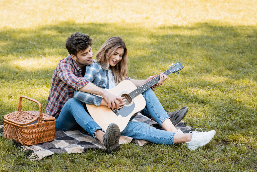 Kareli battaniyede oturan ve erkek arkadaşıyla akustik gitar çalan bir kadın.  - Fotoğraf, Görsel