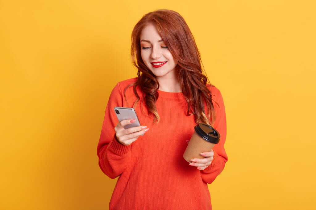Adorable chica con suéter naranja posando aislado sobre fondo amarillo, sosteniendo café para llevar y teléfono móvil, mira la pantalla del dispositivo con sonrisa. - Foto, Imagen