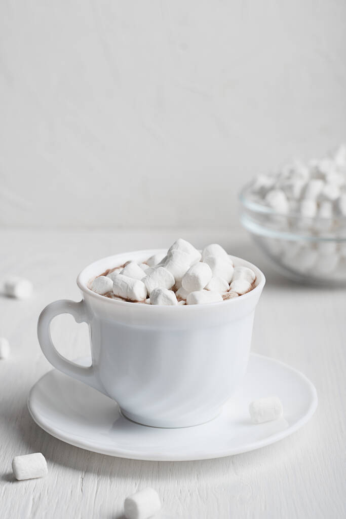 Biały kubek pełen ciepłego słodkiego napoju kakaowego domowej roboty zazwyczaj przygotowywanego w mroźne zimowe i jesienne sesje serwowane na talerzu z miską pianek na drewnianym tle. Obraz pionowej orientacji - Zdjęcie, obraz