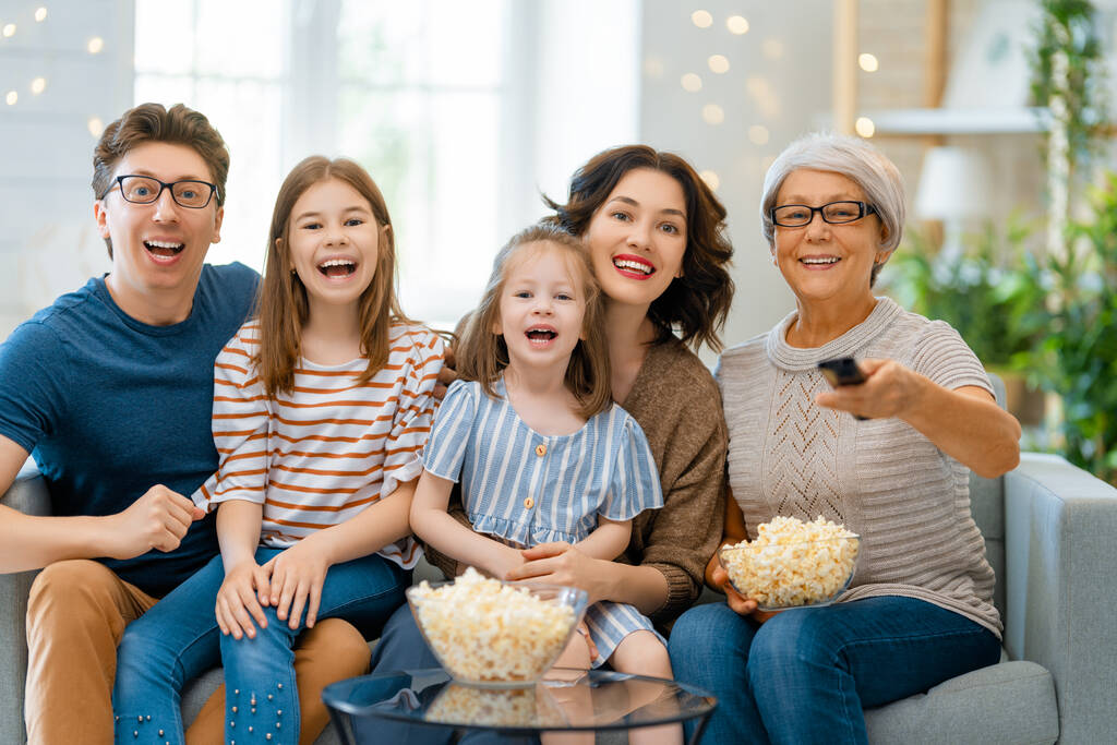 Família feliz assistindo projetor, TV, filmes com pipocas à noite em casa. Avó, mãe, pai e filhas passando tempo juntos. - Foto, Imagem