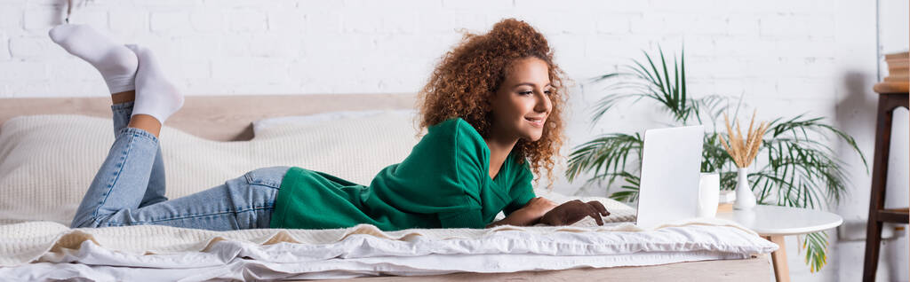 Πανοραμική φωτογραφία της κοκκινομάλλας γυναίκα χρησιμοποιώντας φορητό υπολογιστή, ενώ βρίσκεται στο κρεβάτι  - Φωτογραφία, εικόνα