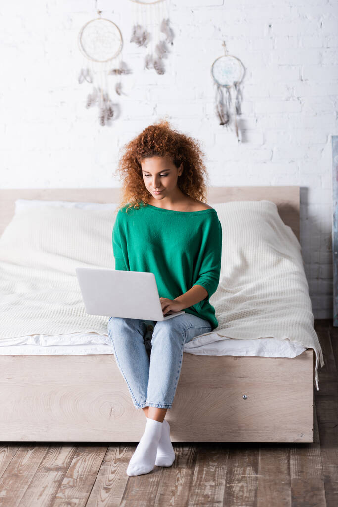 Rothaarige Frau in Jeans benutzt Laptop, während sie auf dem Bett sitzt  - Foto, Bild