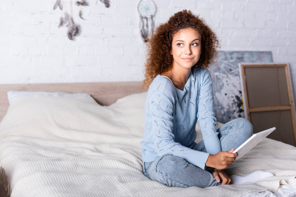 Mujer pelirroja en suéter y jeans sosteniendo tableta digital en la cama  - Foto, imagen