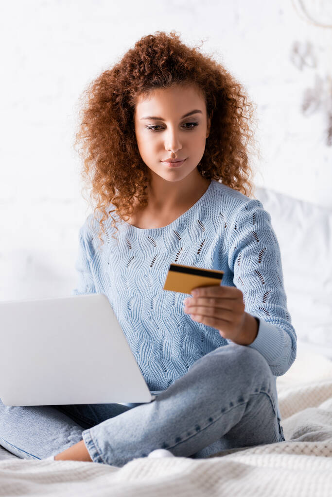Επιλεκτική εστίαση του σγουρά γυναίκα κοιτάζοντας την πιστωτική κάρτα, ενώ κρατώντας φορητό υπολογιστή στο υπνοδωμάτιο  - Φωτογραφία, εικόνα