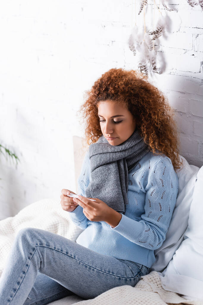 Mulher encaracolada em cachecol e suéter segurando termômetro na cama  - Foto, Imagem