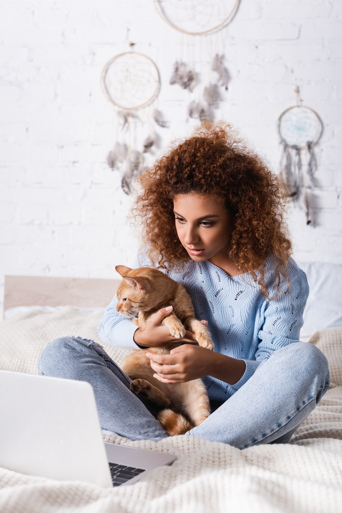 Selektywne skupienie na rudej kobiecie trzymającej kota w pobliżu laptopa na łóżku  - Zdjęcie, obraz
