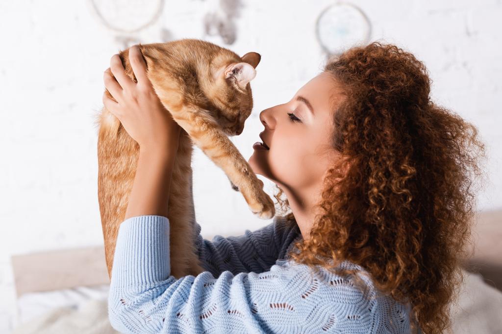 Vista lateral de mujer pelirroja sosteniendo gato en casa  - Foto, imagen
