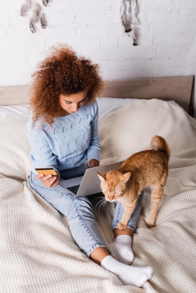 krullende vrouw met behulp van laptop en creditcard in de buurt van gember kat in slaapkamer  - Foto, afbeelding