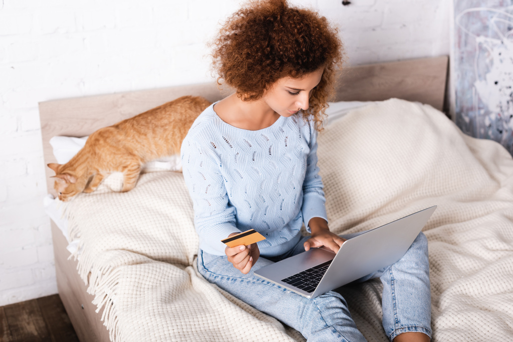 Επιλεκτική εστίαση της νεαρής γυναίκας που κατέχει πιστωτική κάρτα και τη χρήση φορητού υπολογιστή κοντά σε tabby γάτα στο κρεβάτι  - Φωτογραφία, εικόνα