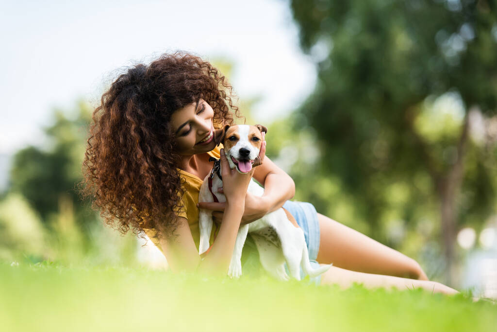 Selektiver Fokus der fröhlichen Frau, die Jack Russell Terrier Hund umarmt, während sie auf dem Rasen liegt - Foto, Bild