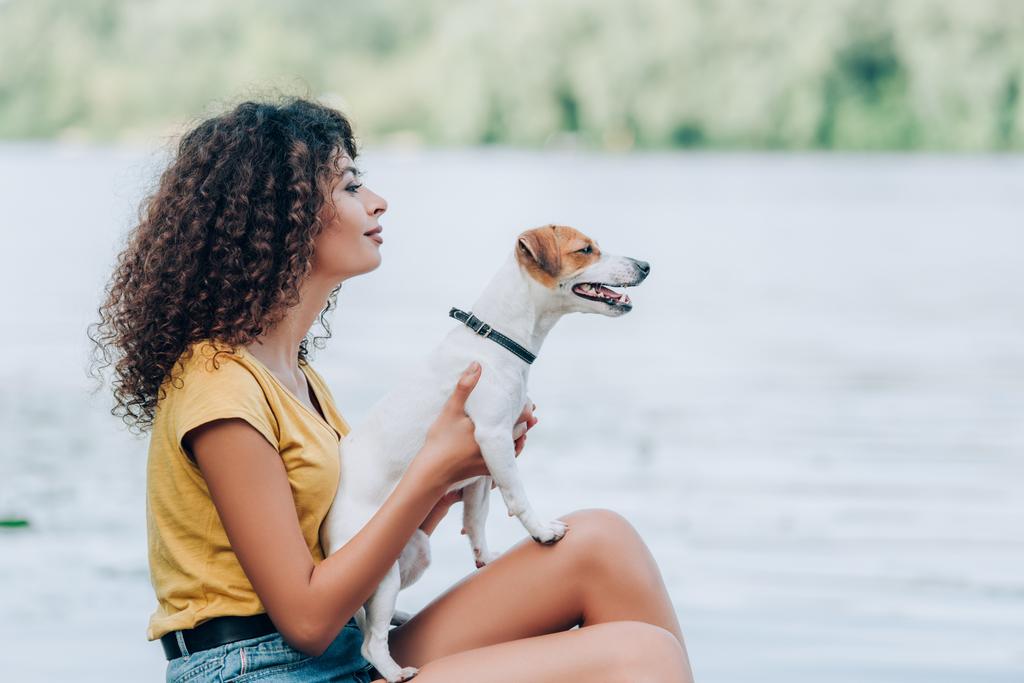 vista lateral de la mujer rizada en traje de verano sosteniendo gato russell terrier perro y mirando hacia otro lado mientras está sentado cerca del lago - Foto, imagen