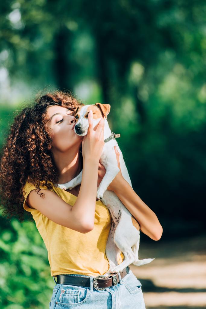 νεαρή γυναίκα με καλοκαιρινά ρούχα φιλάει τον Τζακ Ράσελ Τεριέ σκύλο στο πάρκο - Φωτογραφία, εικόνα