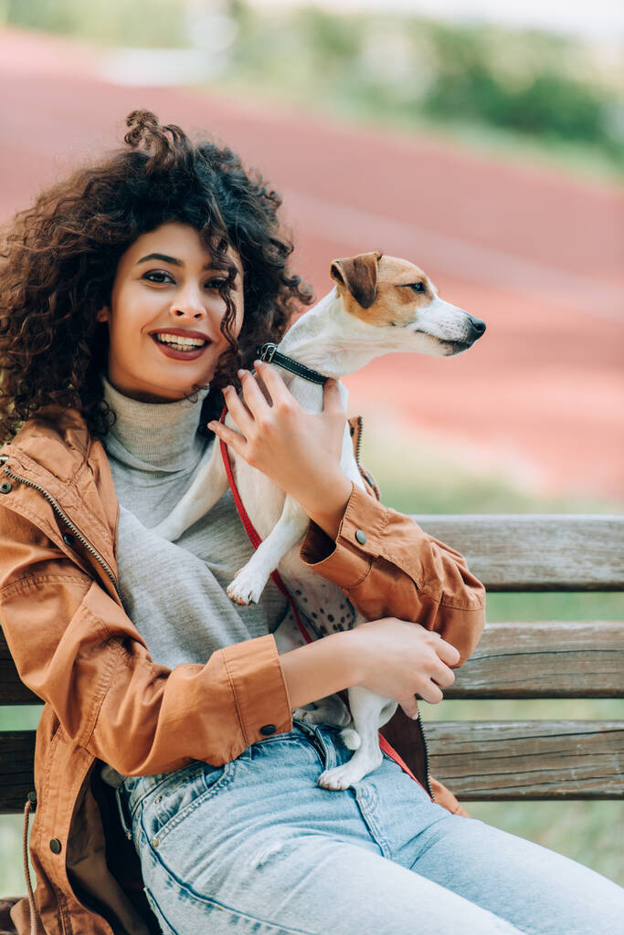 Kıvırcık kadın sonbahar kıyafetiyle kameraya bakarken parkta bankta Jack Russell Terrier 'a sarılıyor. - Fotoğraf, Görsel