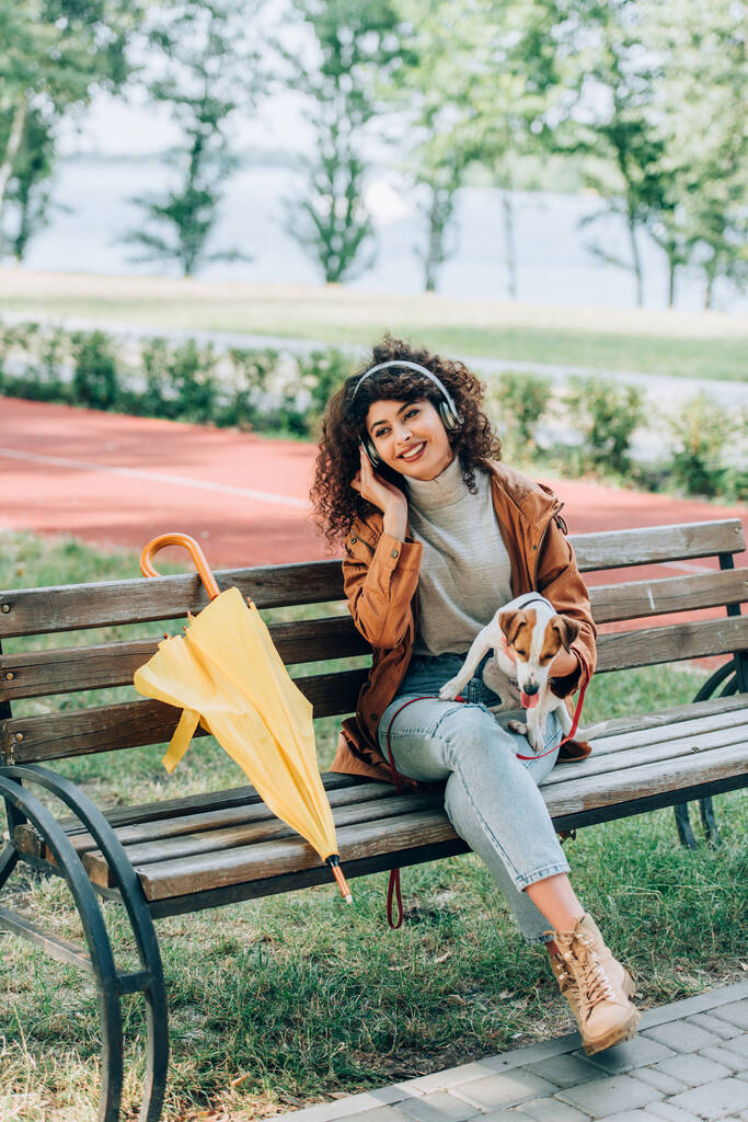 κομψή γυναίκα σε ασύρματα ακουστικά κρατώντας jack Russell terrier σκυλί, ενώ κάθεται στον πάγκο κοντά στο κίτρινο ομπρέλα - Φωτογραφία, εικόνα