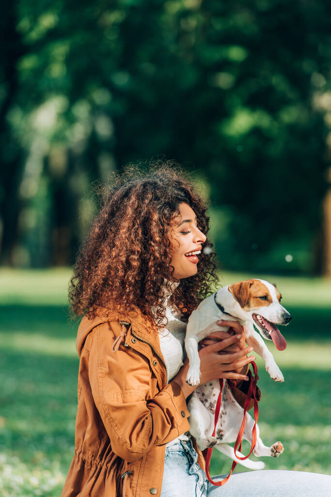 Kędzierzawa kobieta w płaszczu przeciwdeszczowym trzymająca Jacka Russella Terriera ze smycz w parku  - Zdjęcie, obraz