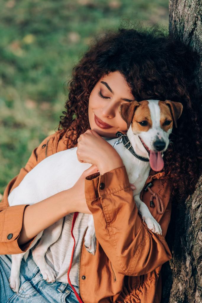 Young woman in raincoat hugging jack russell terrier near tree  - Photo, Image