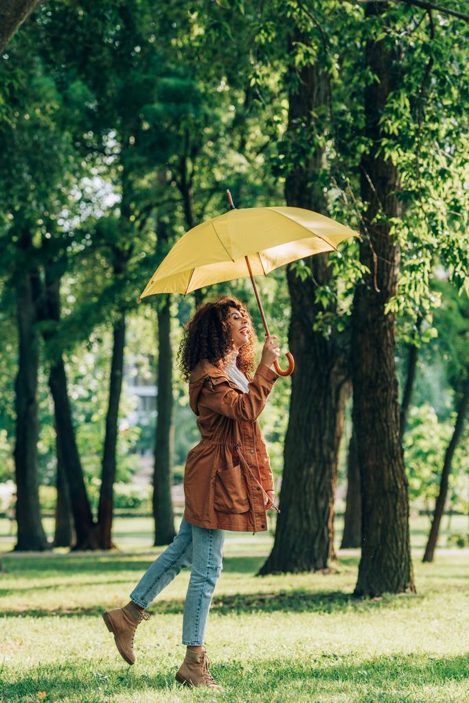 Vista lateral da mulher encaracolada em capa de chuva e jeans segurando guarda-chuva no parque  - Foto, Imagem