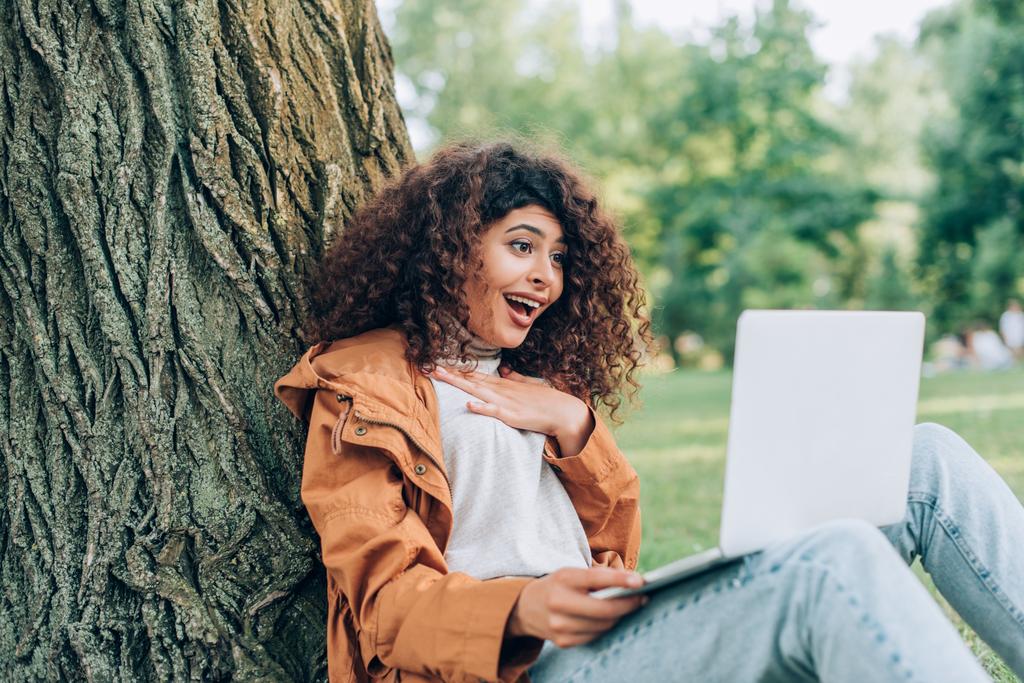 公園の木の近くにノートパソコンを使用して興奮した女性の選択的な焦点  - 写真・画像