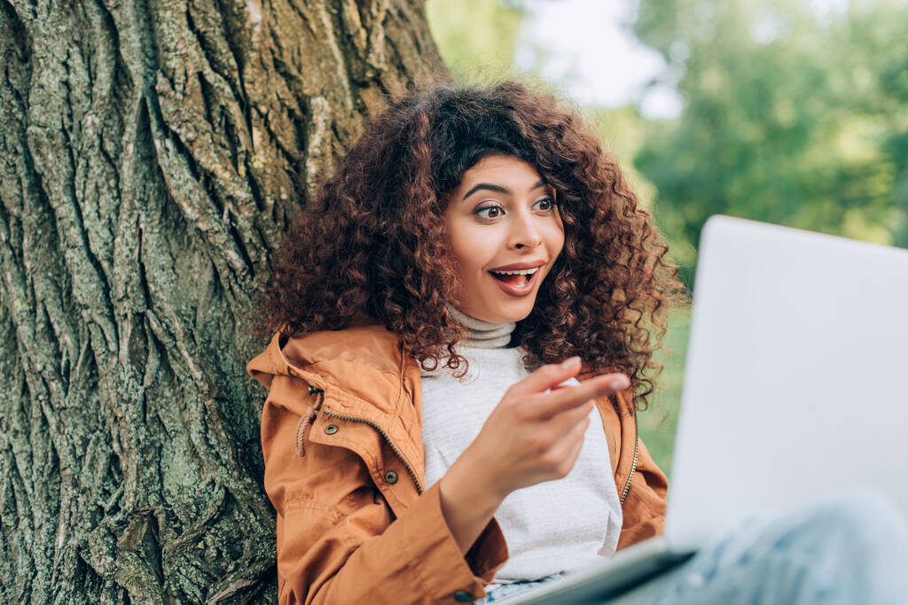 Mise au point sélective d'un pigiste excité pointant du doigt un ordinateur portable près d'un arbre dans un parc  - Photo, image