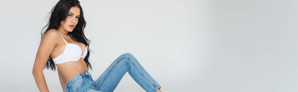 panoramic crop of sexy and brunette woman in jeans and bra isolated on grey - Photo, Image
