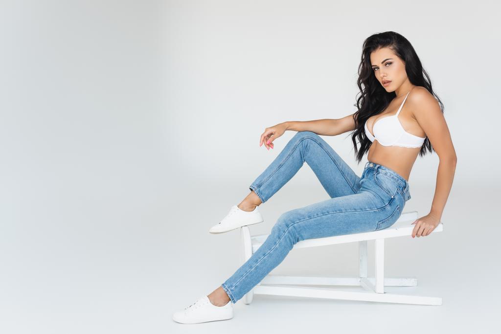 brunette woman in denim jeans and bra sitting on stool on grey - Photo, Image