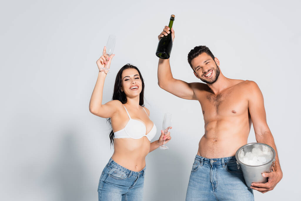 bez koszulki mężczyzna trzyma wiadro lodu i butelkę szampana w pobliżu seksownej kobiety w staniku stojąc w okularach na szarości - Zdjęcie, obraz
