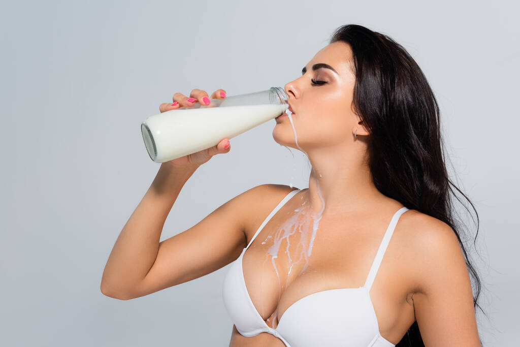 sexy woman in bra holding bottle and drinking milk isolated on grey - Photo, Image