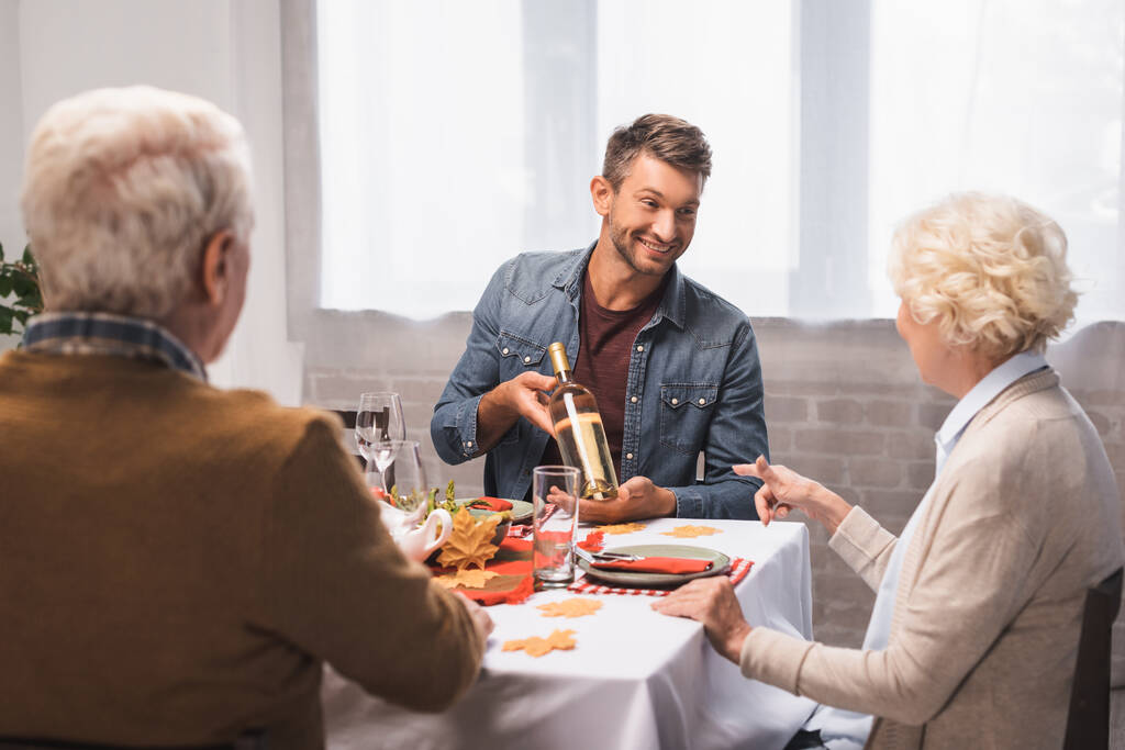 donna anziana che punta con il dito alla bottiglia di vino nelle mani del figlio gioioso durante la cena del Ringraziamento - Foto, immagini