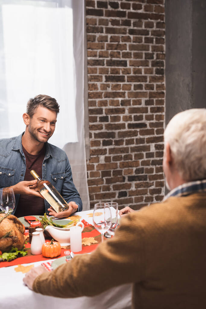 selectieve focus van vrolijke man tonen fles witte wijn aan senior vader op dankzegging diner - Foto, afbeelding