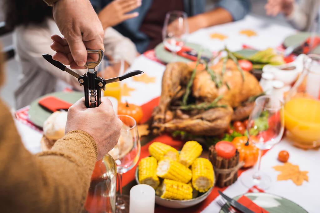 gedeeltelijk zicht op senior man openen fles witte wijn met kurkentrekker tijdens Thanksgiving diner met multi-etnische familie - Foto, afbeelding