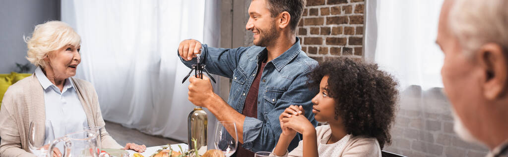 panoramiczna koncepcja człowieka otwierającego butelkę białego wina podczas kolacji dziękczynnej z wielokulturową rodziną - Zdjęcie, obraz
