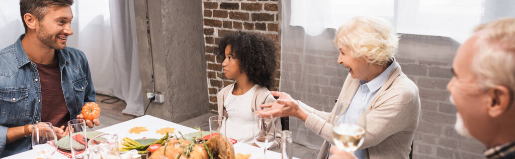 panoramatický záběr vzrušené ženy gestikulující během večeře na Díkůvzdání s multikulturní rodinou - Fotografie, Obrázek
