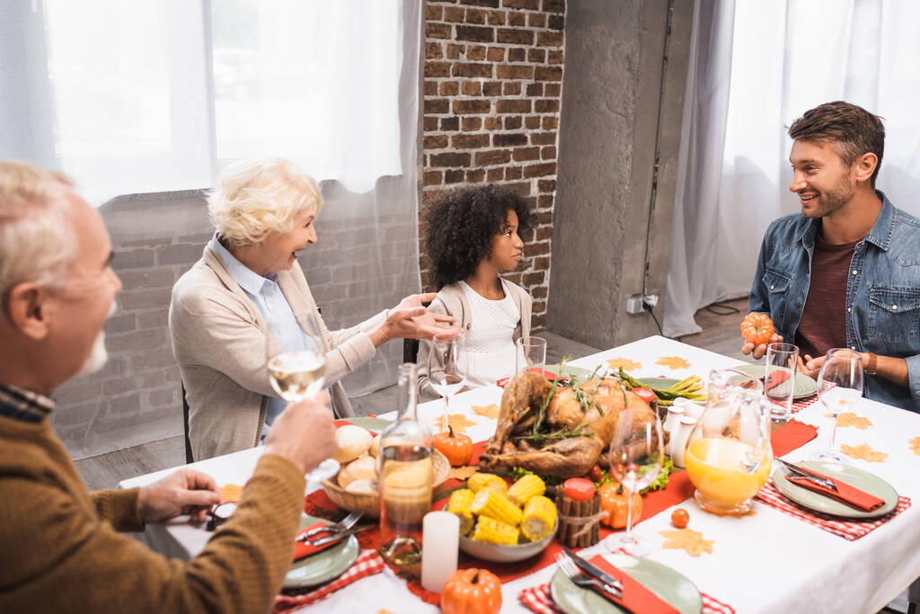 пожилая женщина жестикулирует, разговаривая с мультикультурной семьей во время ужина в честь Дня благодарения - Фото, изображение