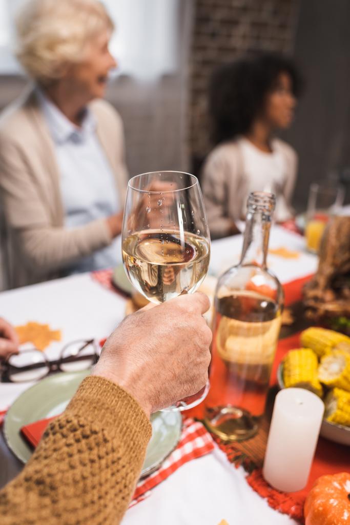 senior man holding glass of white wine while celebrating thanksgiving with multicultural family - Photo, Image