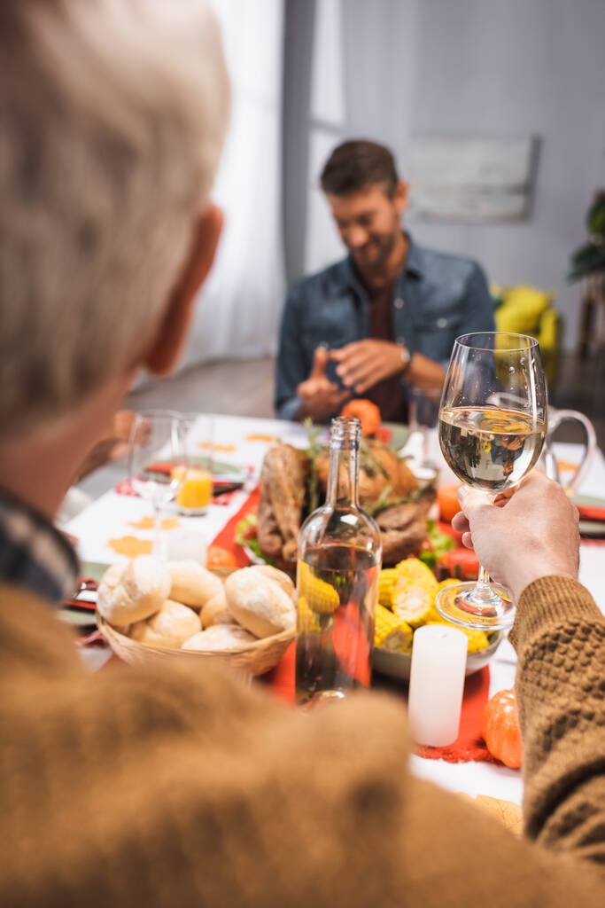επιλεκτική εστίαση του ηλικιωμένου άνδρα κρατώντας ποτήρι λευκό κρασί κατά τη διάρκεια των Ευχαριστιών δείπνο με την οικογένεια - Φωτογραφία, εικόνα