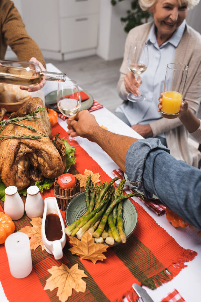 обрезанный вид человека, наливающего белое вино во время празднования Дня благодарения с мультикультурной семьей - Фото, изображение