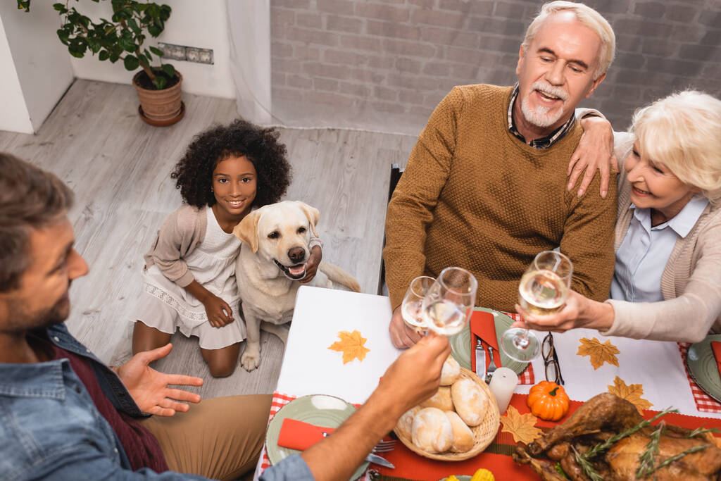 high angle view of african american girl and golden retriever near family clinking wine glasses during thanksgiving dinner - Photo, Image
