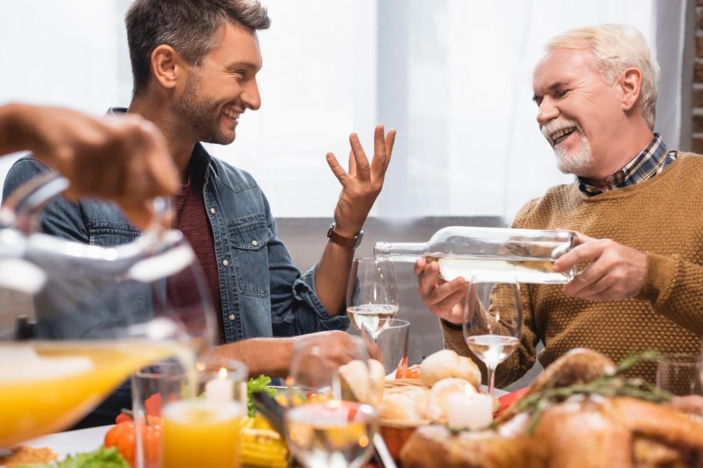 joyful man gesturing while talking to father pouring white wine during thanksgiving dinner - Photo, Image