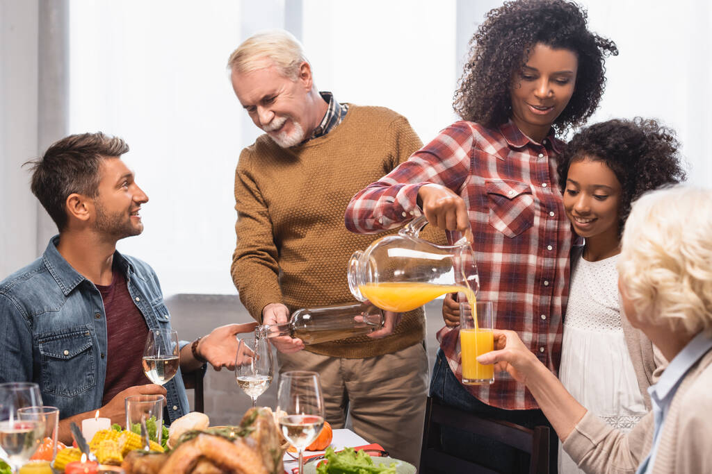 afrikanisch-amerikanische Frau und älterer Mann gießen Orangensaft und Weißwein während des Dankeschön-Dinners mit multikultureller Familie - Foto, Bild