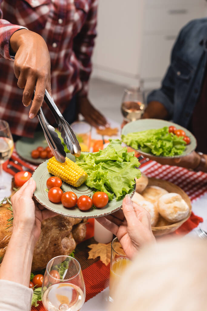 częściowy widok starszej kobiety trzymającej talerz z kukurydzą, sałatą i pomidorami wiśniowymi podczas kolacji dziękczynnej z wieloetniczną rodziną - Zdjęcie, obraz