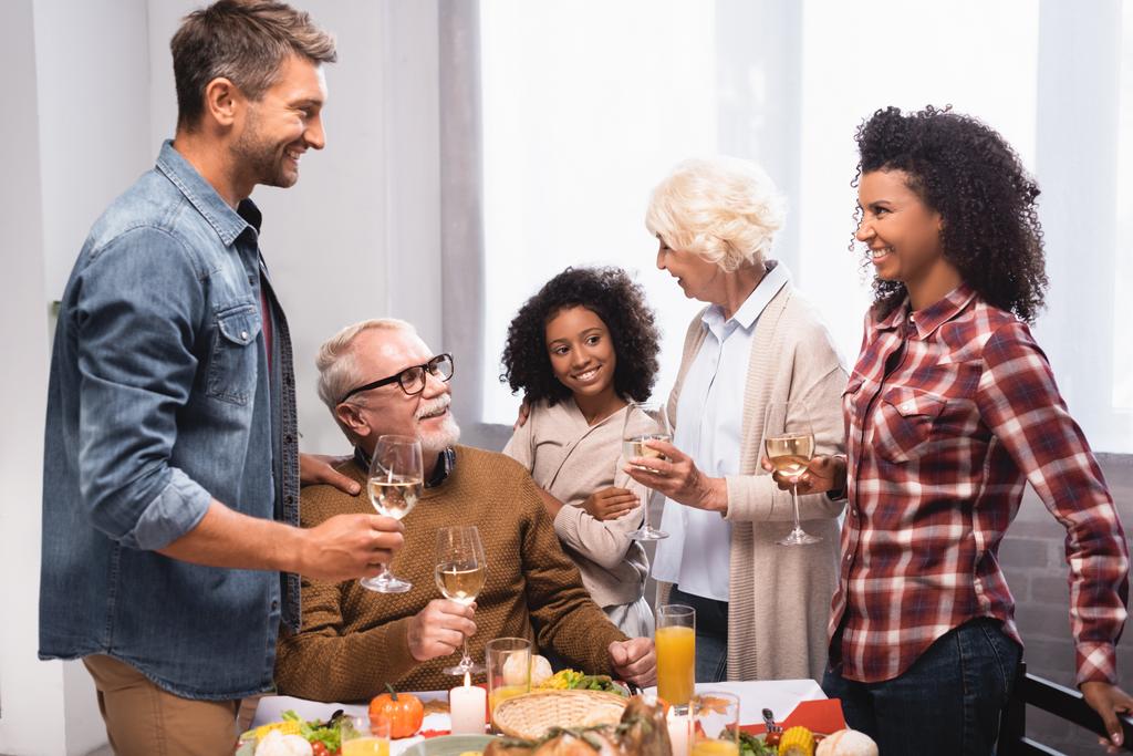 família multiétnica animado falando enquanto segurando copos de vinho na mesa servida com jantar de ação de graças - Foto, Imagem