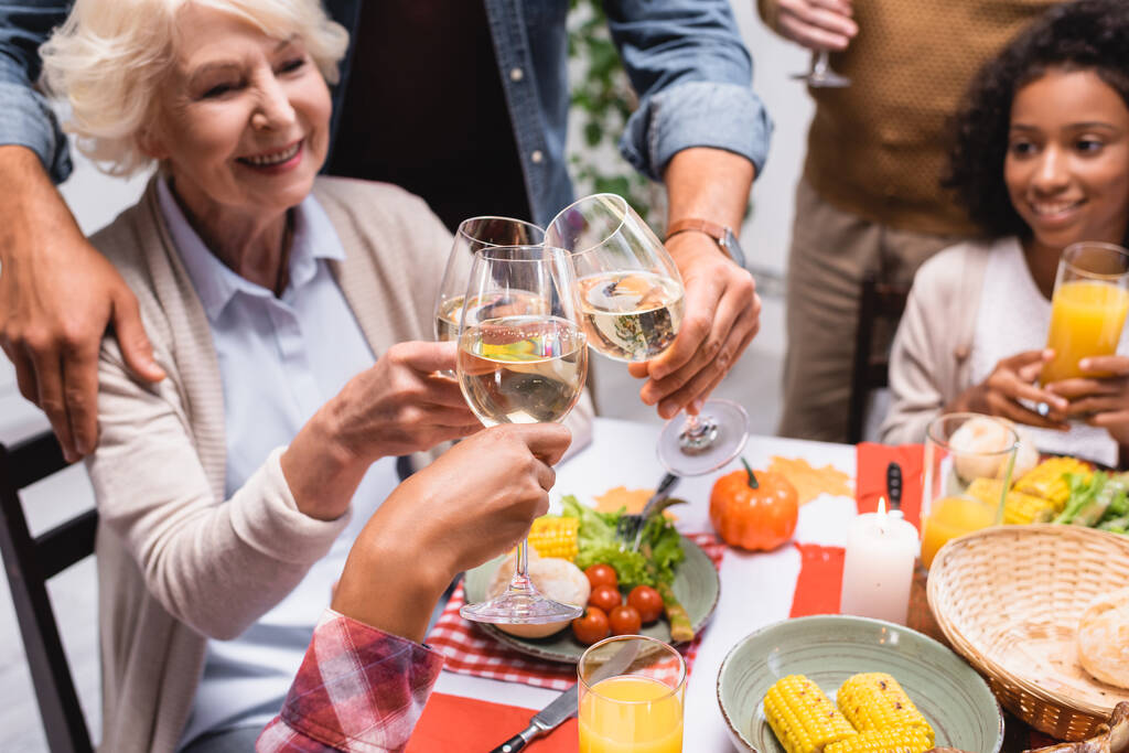 selektywne skupienie seniorki brzęczącej kieliszkami wina z wielokulturową rodziną podczas kolacji dziękczynnej - Zdjęcie, obraz