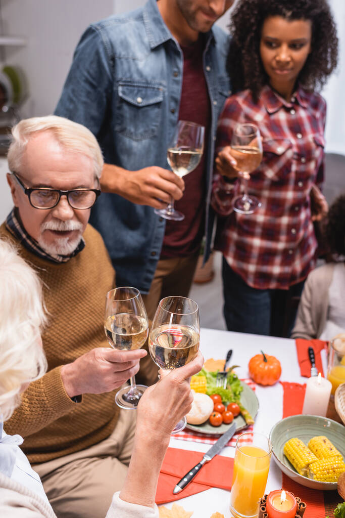 Selective focus of elderly woman clinking wine with man during thanksgiving celebration with multiethnic family  - Photo, Image